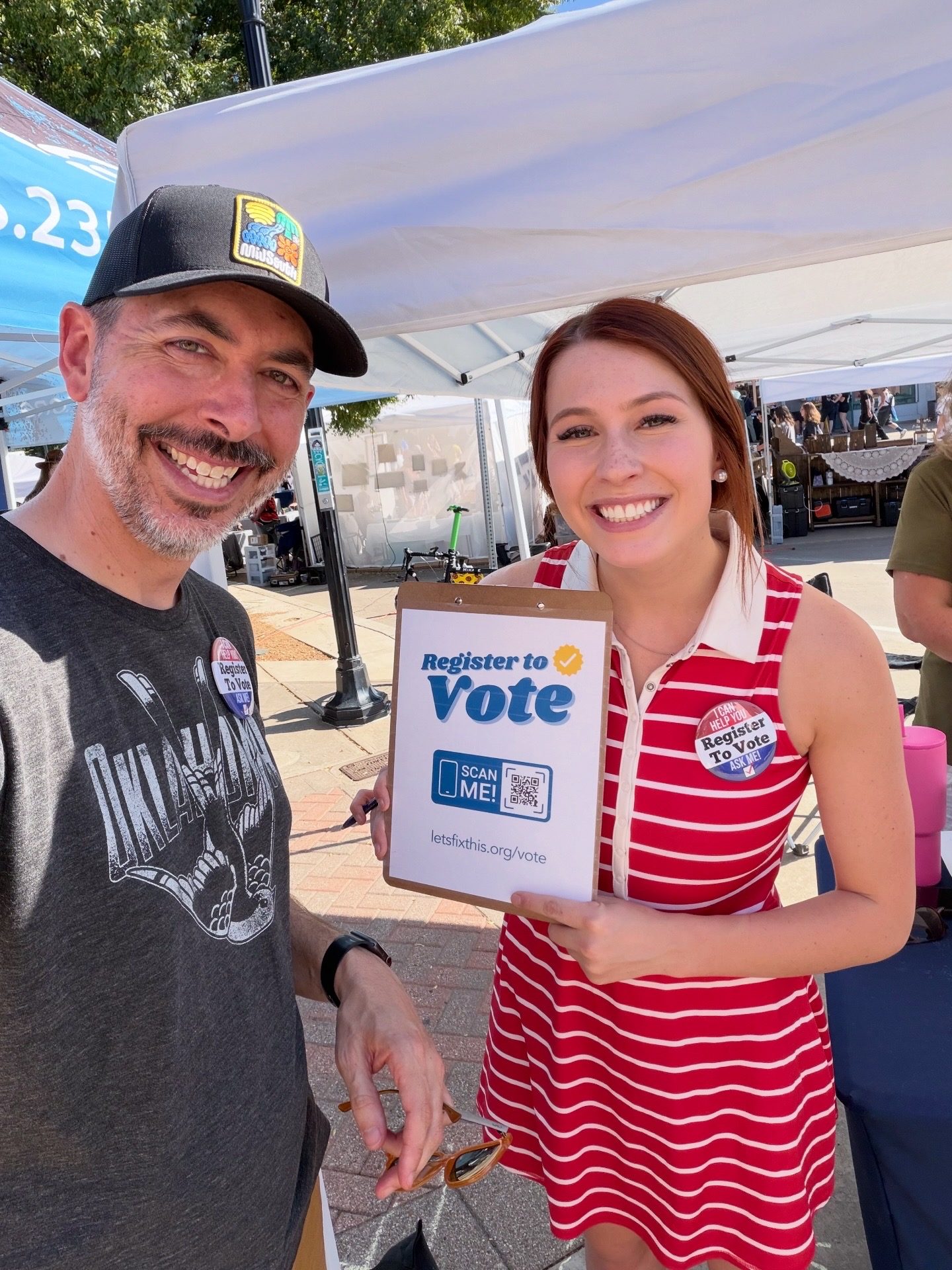 Two smiling volunteers hold a Register to Vote clipboard with a QR code at an outdoor market.
