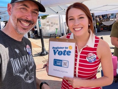 Two smiling volunteers hold a Register to Vote clipboard with a QR code at an outdoor market.