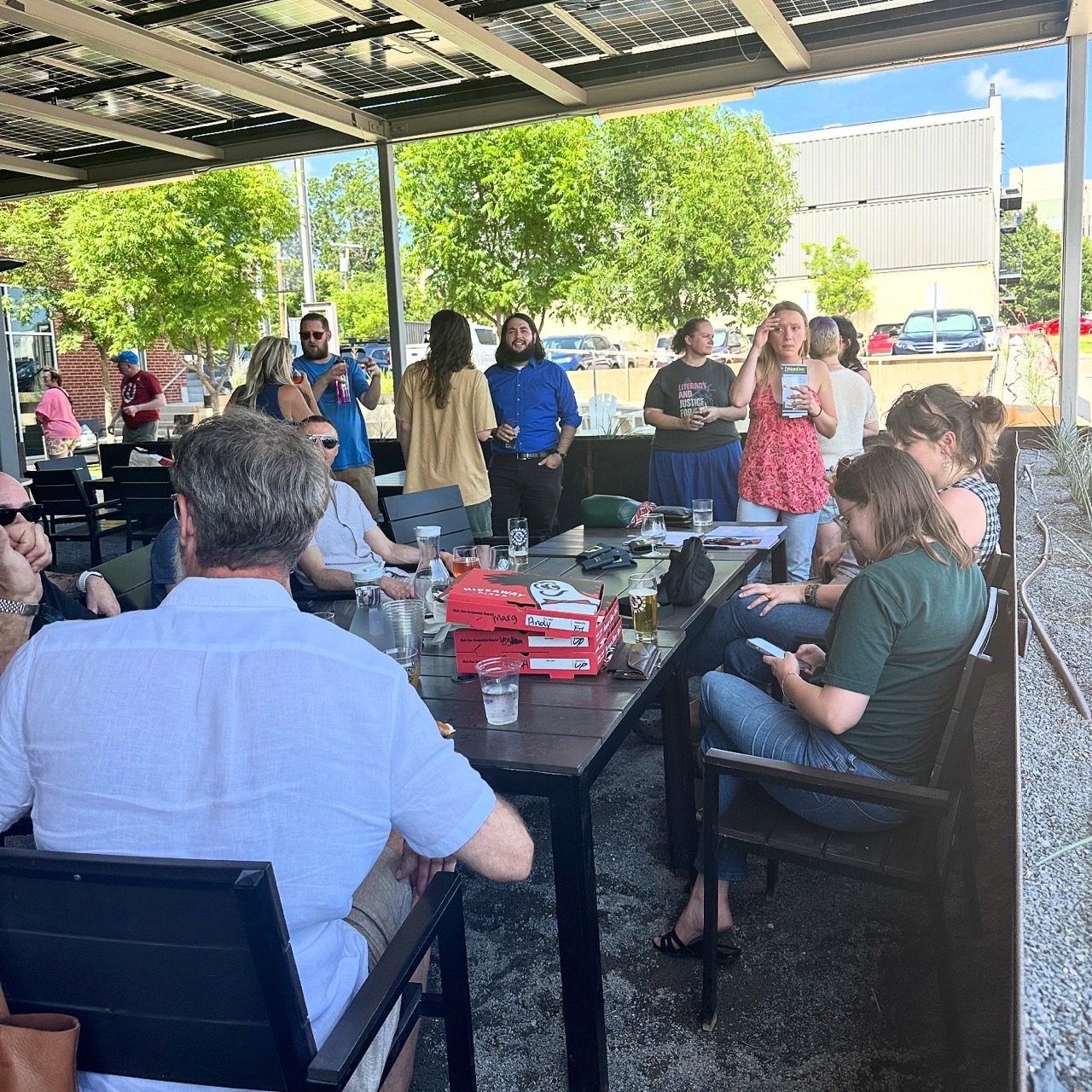 Guests sit around patio tables with pizza boxes while others stand and talk.