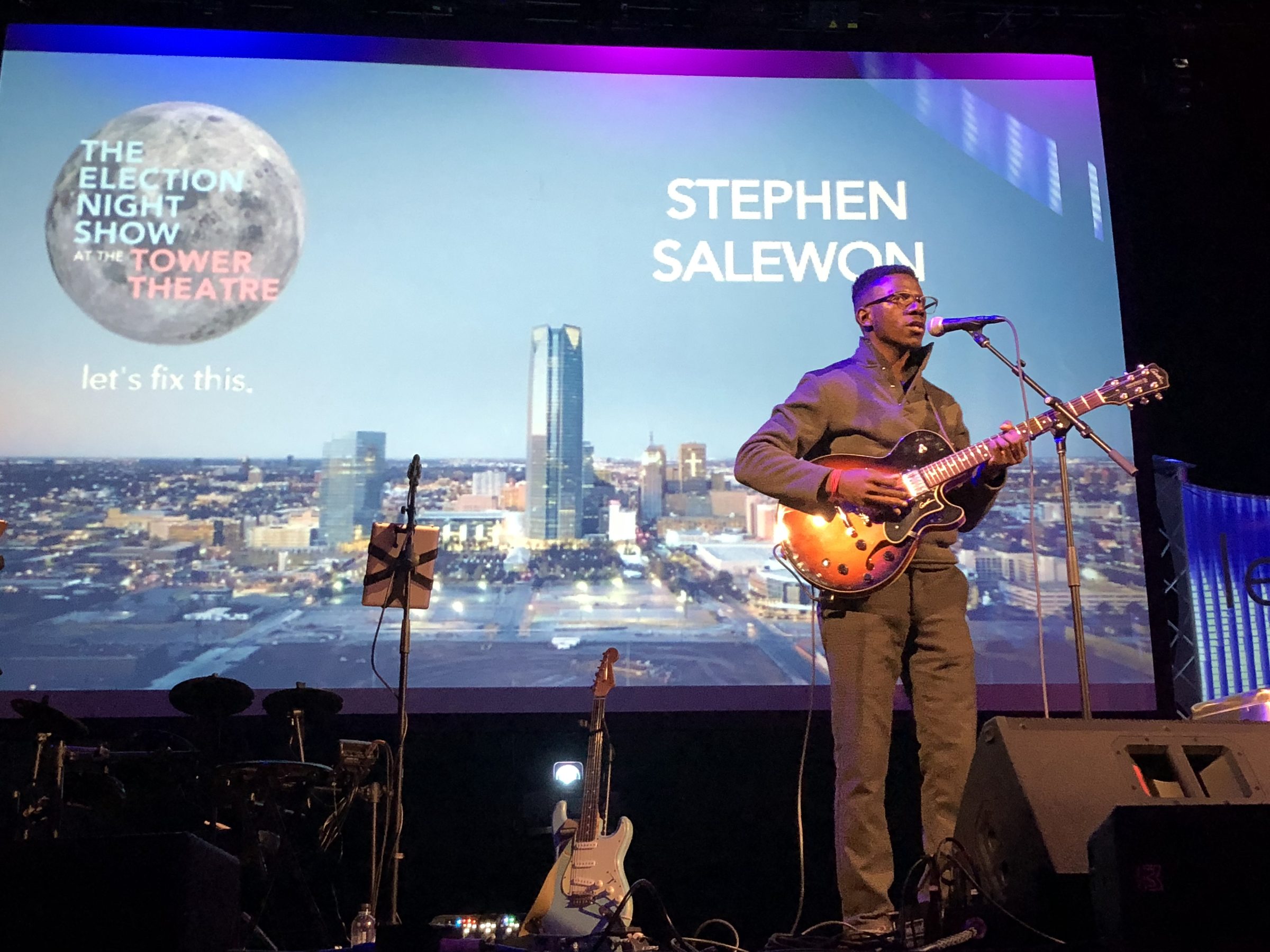 A guitarist performs on stage in front of a screen showing The Election Night Show at the Tower Theatre graphic.