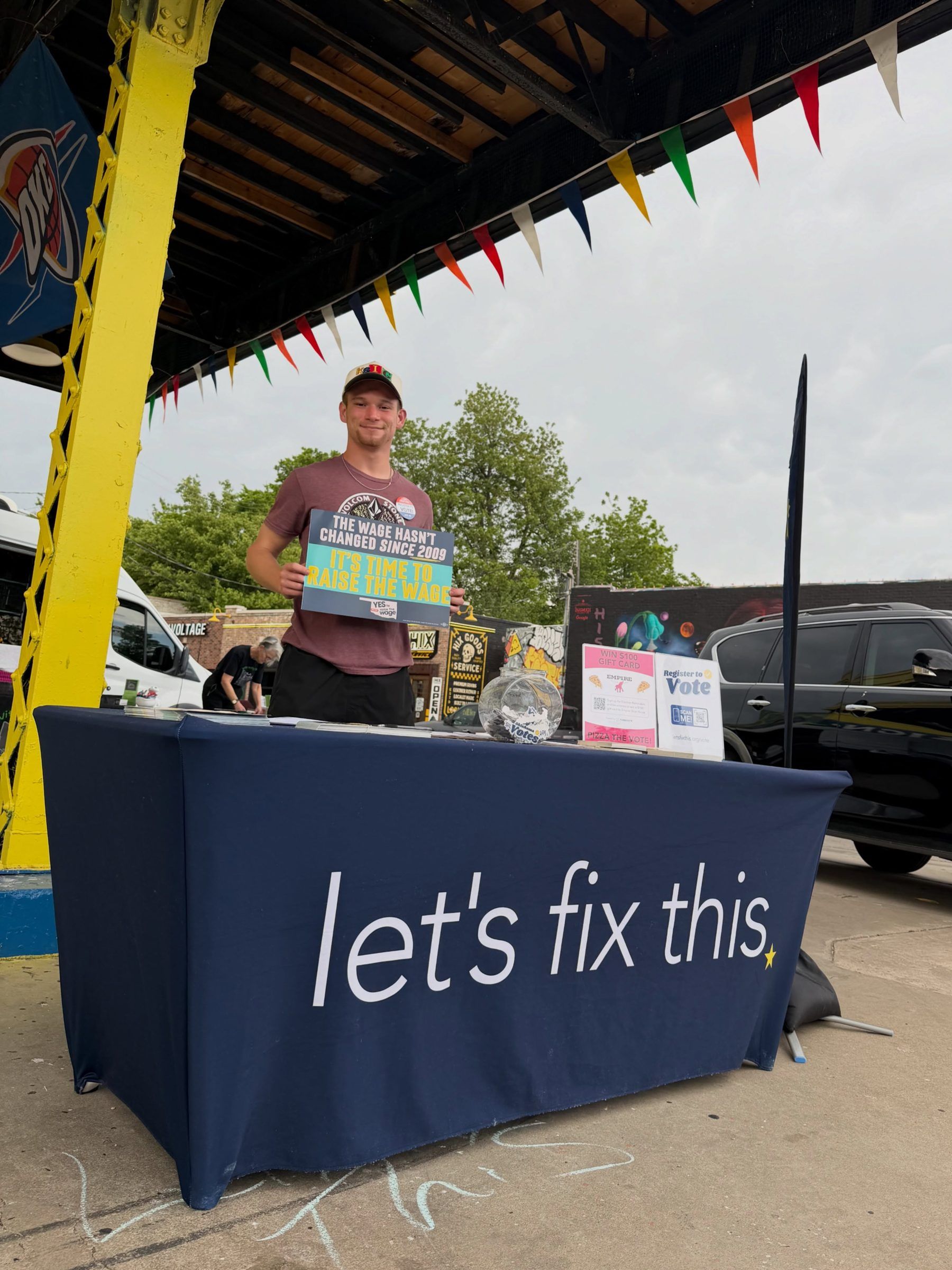 A volunteer holds up a clipboard behind a Let's Fix This table at an outdoor street festival.