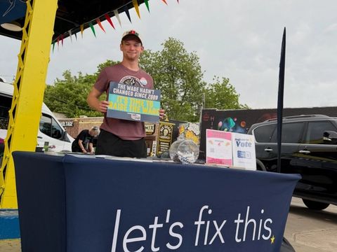 A volunteer holds up a clipboard behind a Let's Fix This table at an outdoor street festival.