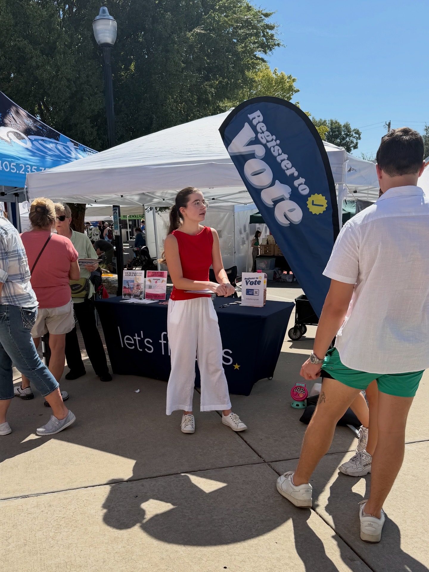 A volunteer stands at a Let's Fix This booth under a Register to Vote flag as festival-goers pass by.