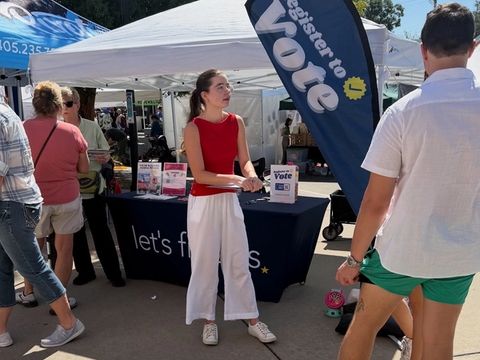 A volunteer stands at a Let's Fix This booth under a Register to Vote flag as festival-goers pass by.