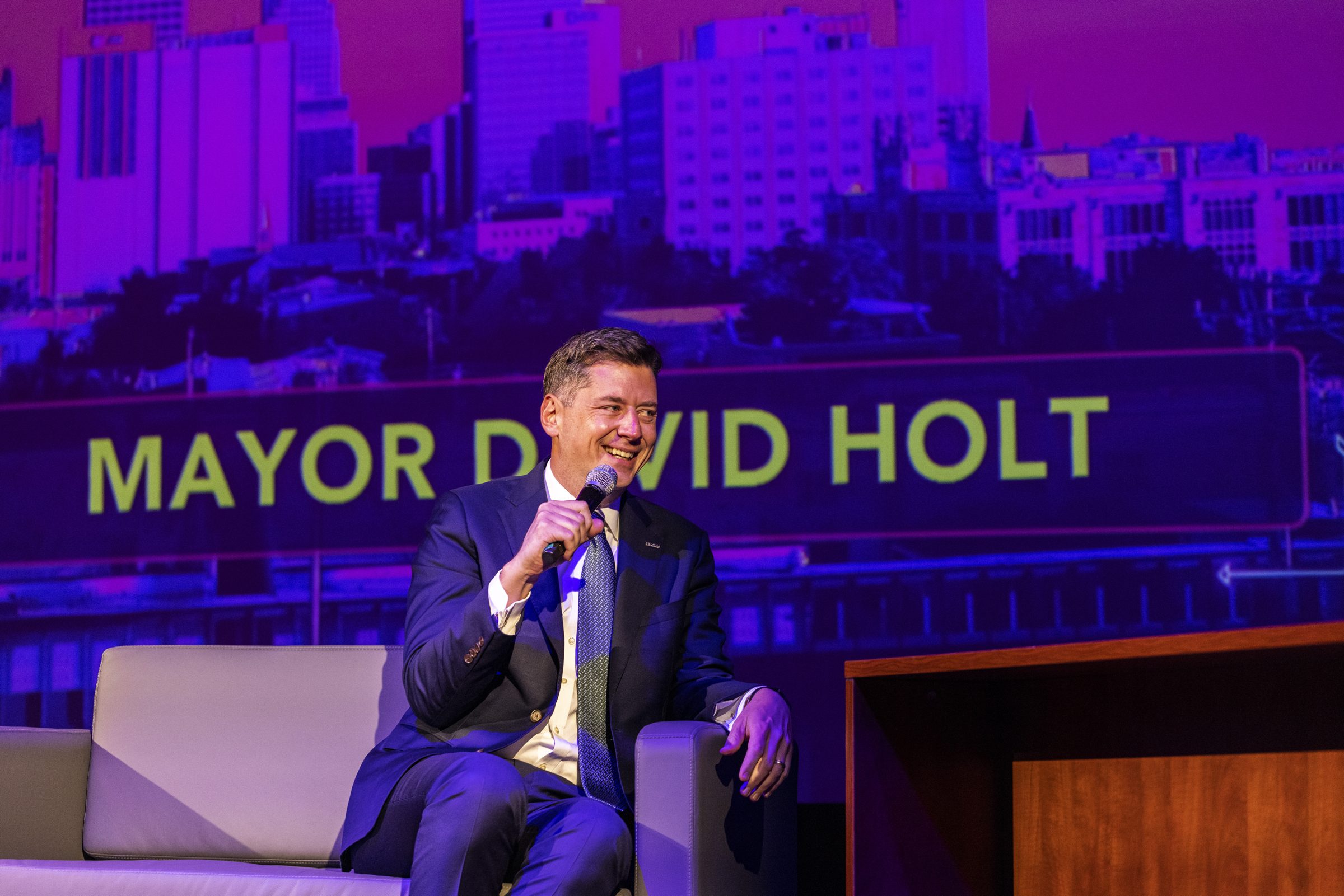A guest in a suit talks with the host on stage, his name projected behind him.