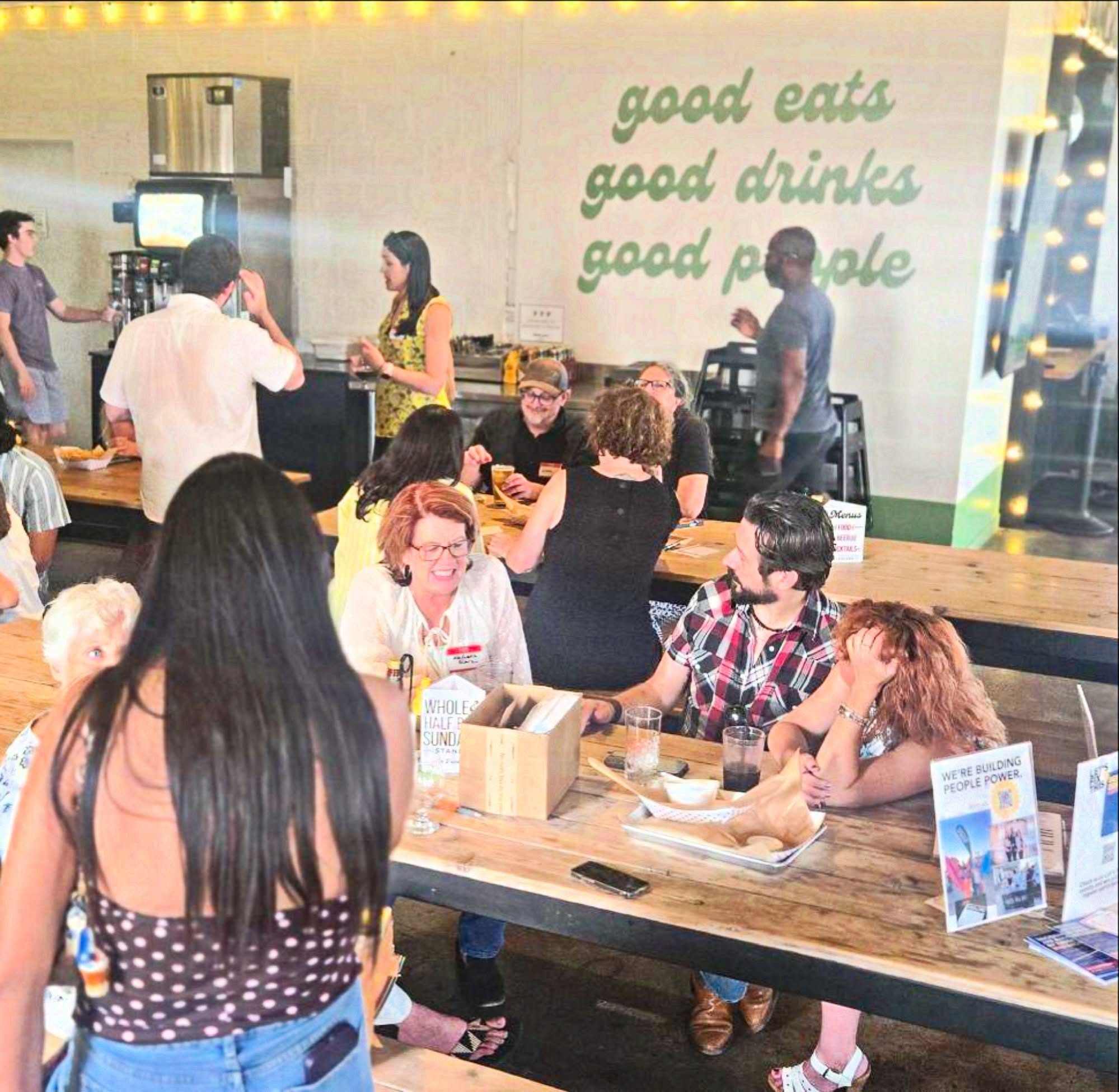 A busy restaurant table at Democracy Drinks in Norman, under a wall sign reading good eats, good drinks, good people.