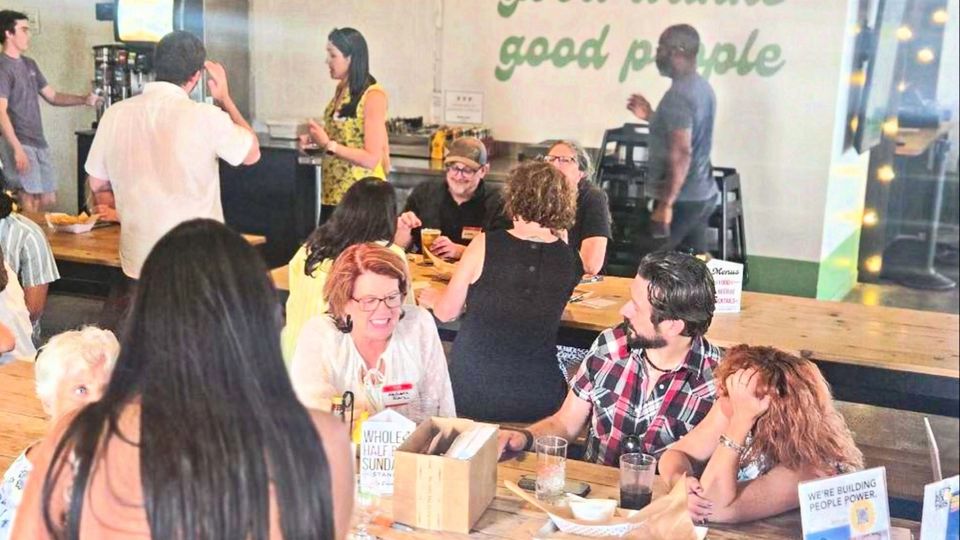 A busy restaurant table at Democracy Drinks in Norman, under a wall sign reading good eats, good drinks, good people.