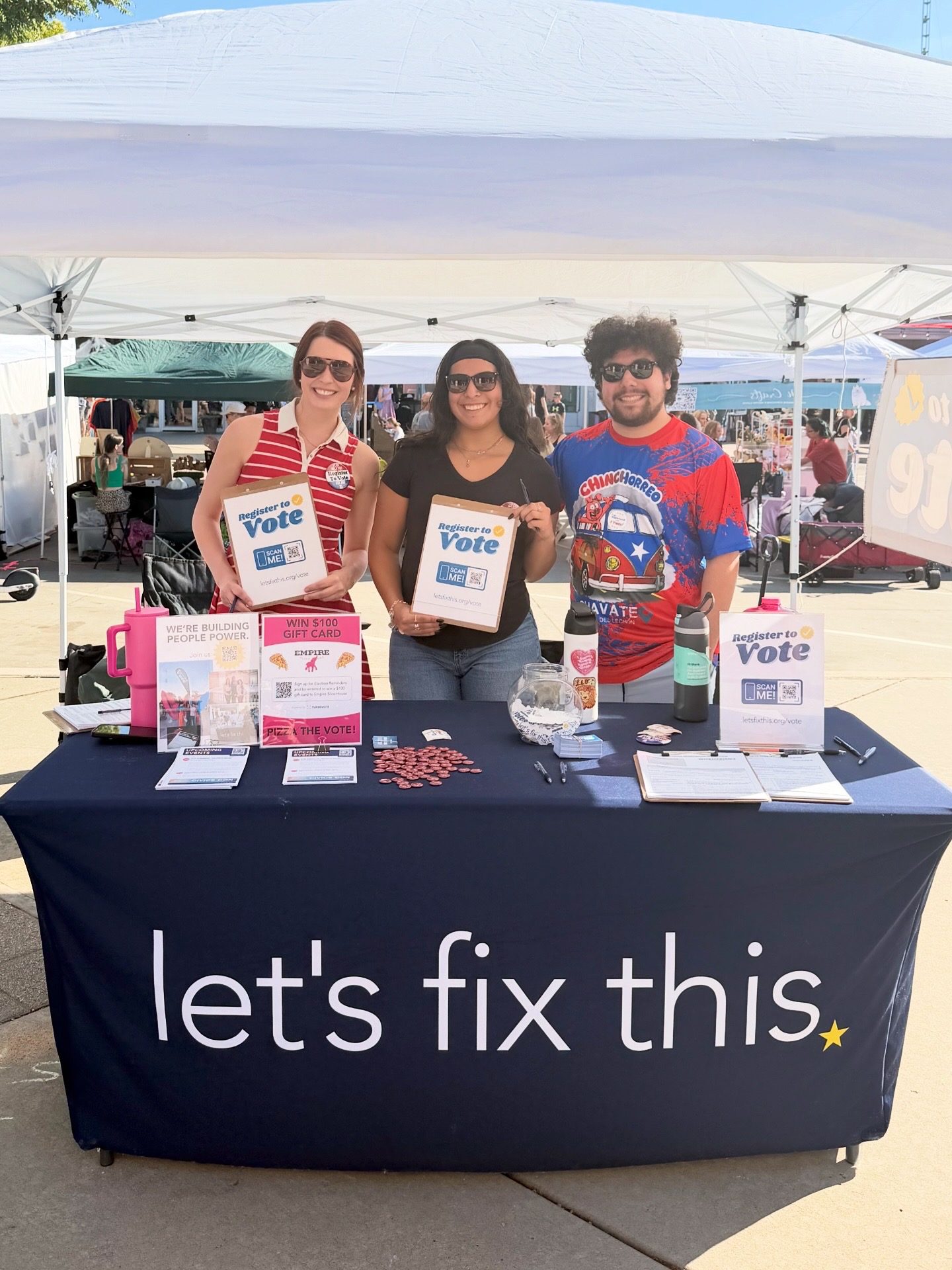 Three volunteers hold register-to-vote signs behind a Let's Fix This table at an outdoor festival.