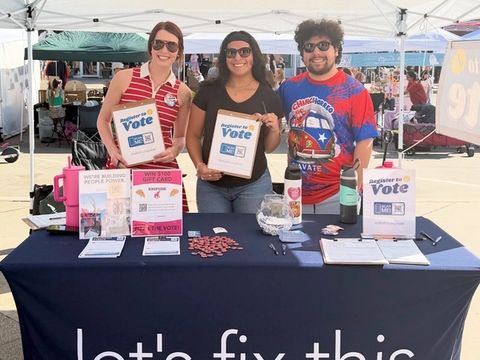 Three volunteers hold register-to-vote signs behind a Let's Fix This table at an outdoor festival.