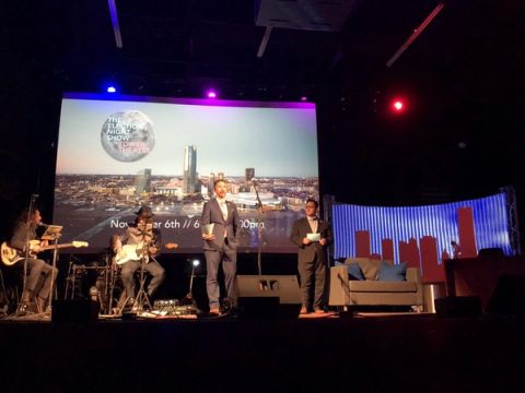 Hosts and musicians share a stage lit in blue and purple, with an Oklahoma City skyline on the big screen.