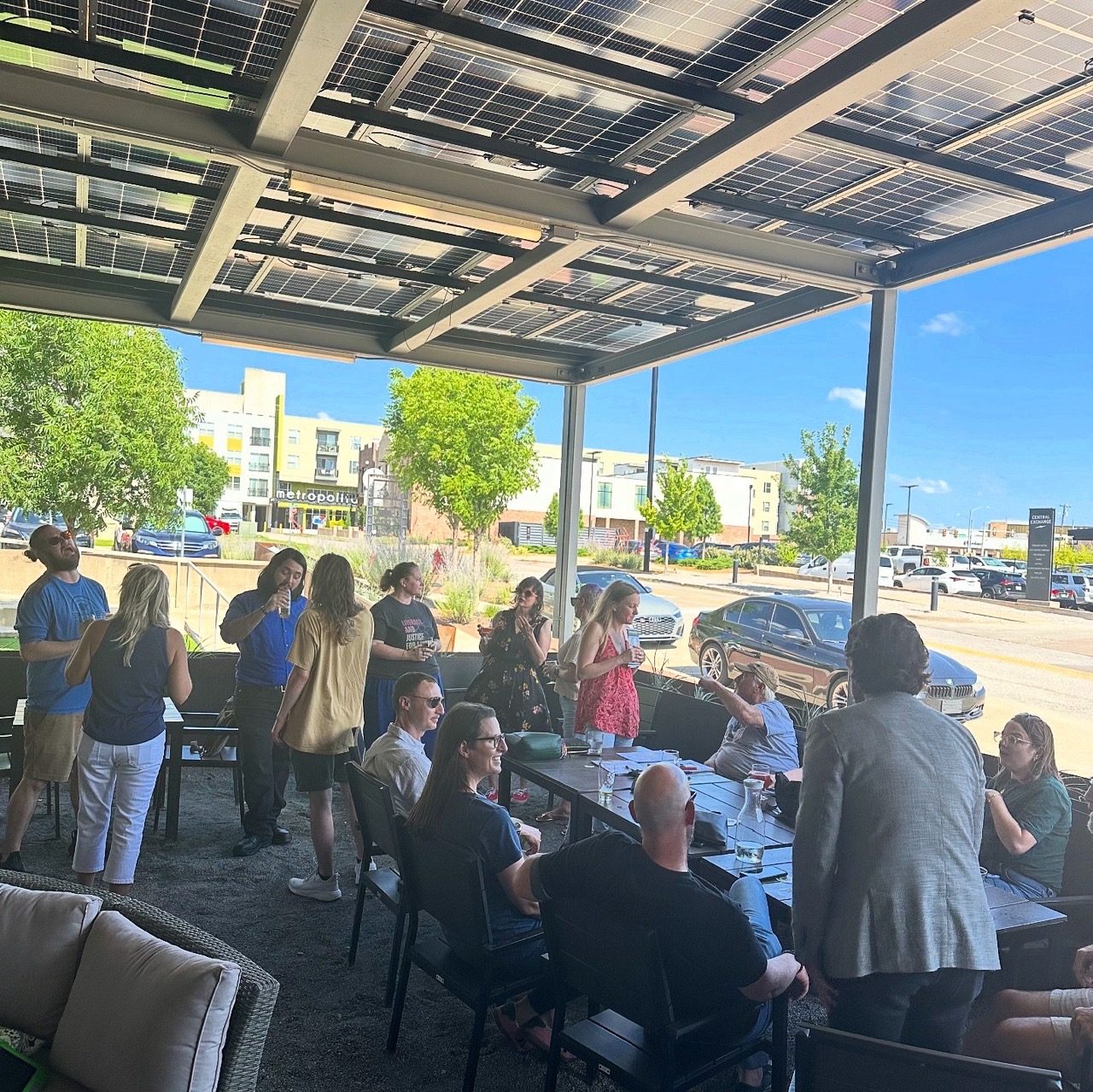 People gather and chat under a solar-panel patio canopy on a sunny afternoon.