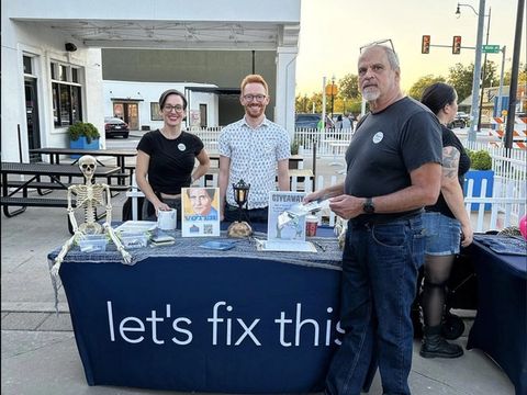 Three volunteers staff a Let's Fix This registration table decorated with a Halloween skeleton.
