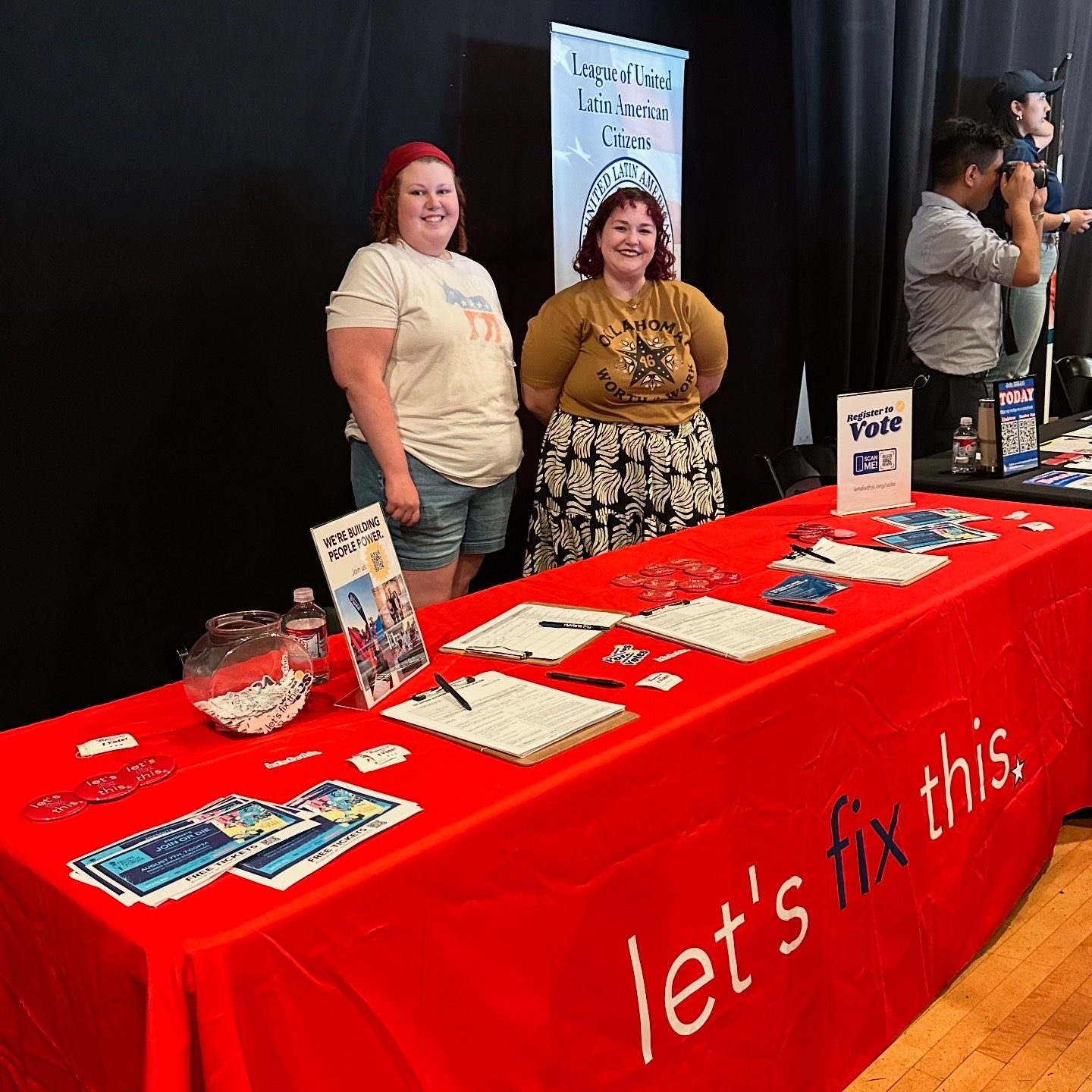 Two volunteers stand behind a red Let's Fix This table with voter registration materials, next to a partner organization's banner.