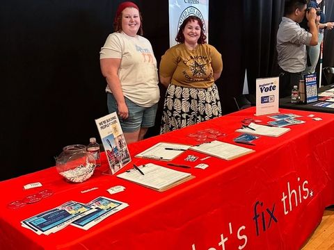 Two volunteers stand behind a red Let's Fix This table with voter registration materials, next to a partner organization's banner.
