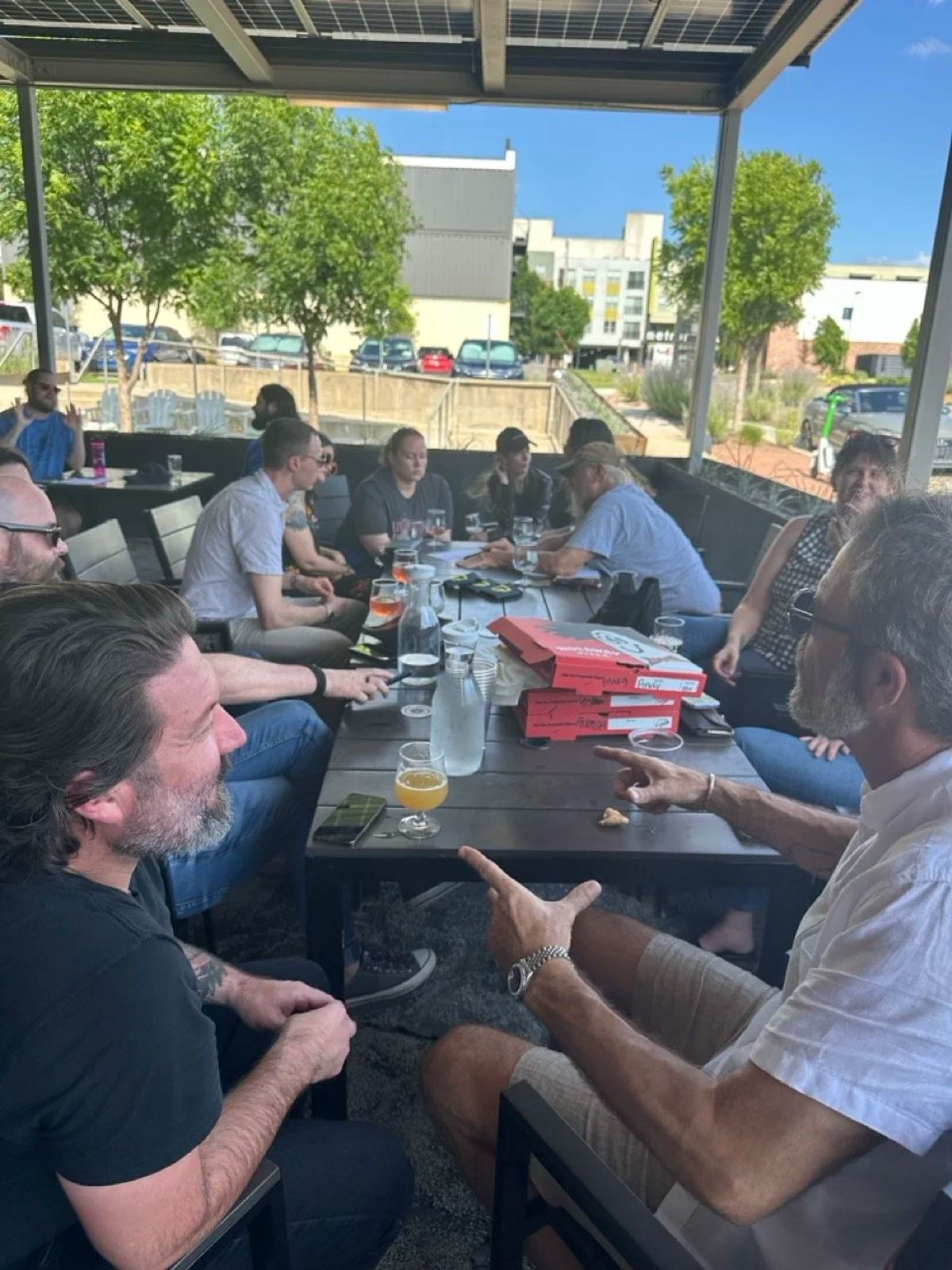 A Democracy Drinks crowd talks around a long patio table on a sunny afternoon.