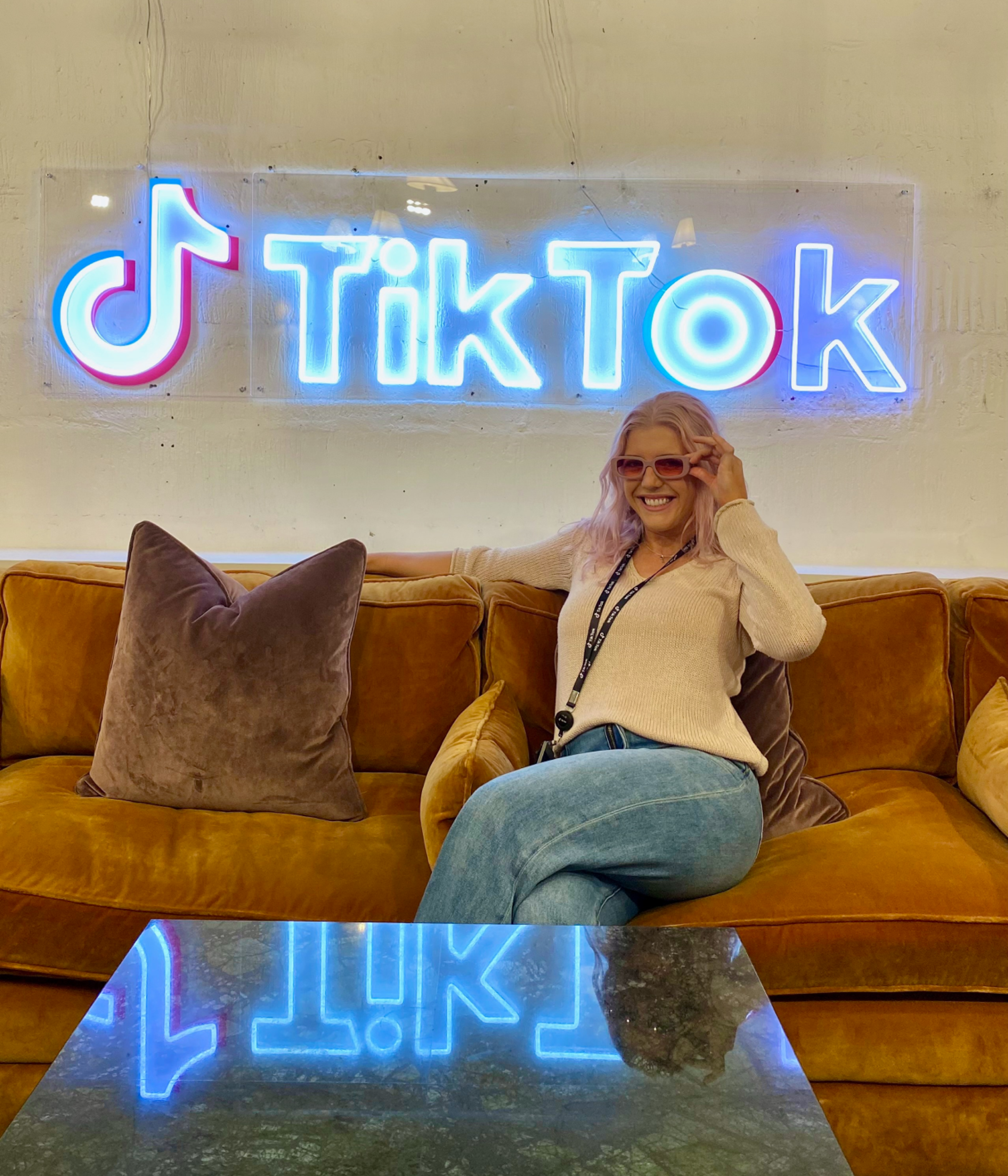 A blond female TikTok employee sits and smiles on a couch with a neon TikTok logo sign on the wall behind her.