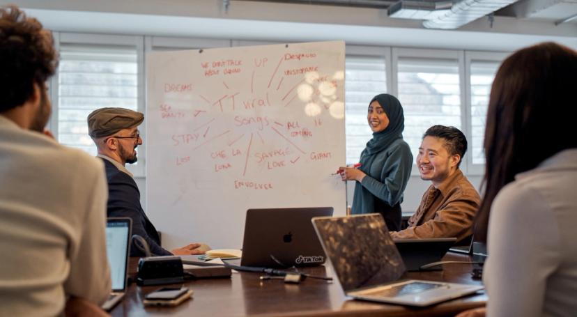 A diverse group of TikTok Ph.D. Interns sit and watch a female colleague, who wears a hijab, as she writes on a white board.