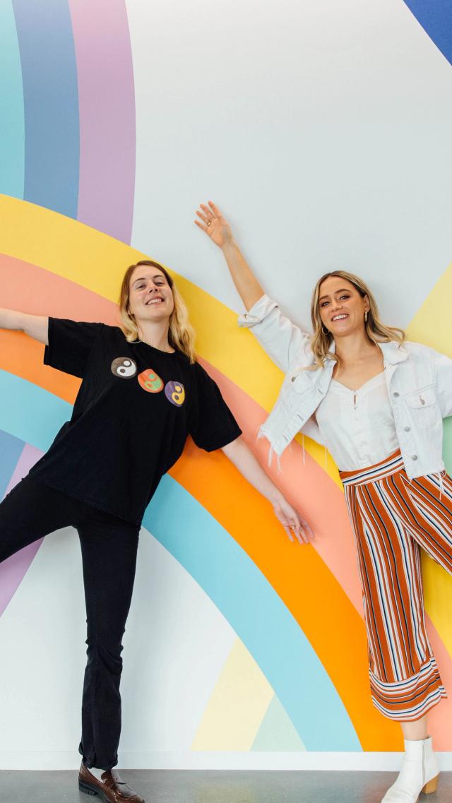 Two young female employees pose in front of a rainbow wall