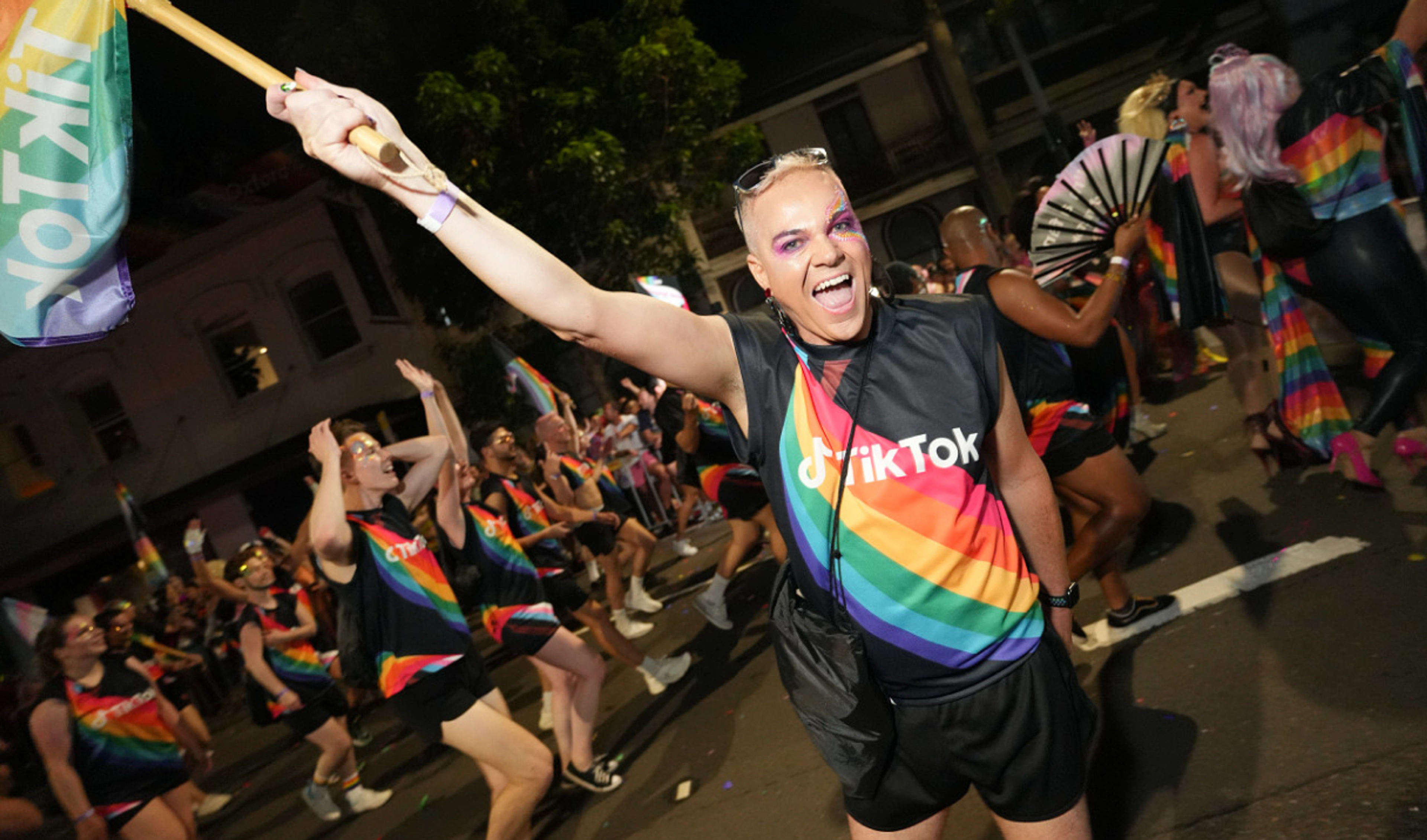 A young man wearing a Tee shirt with the TikTok logo and a rainbow graphic waves a flag at an LGBTQ street parade.