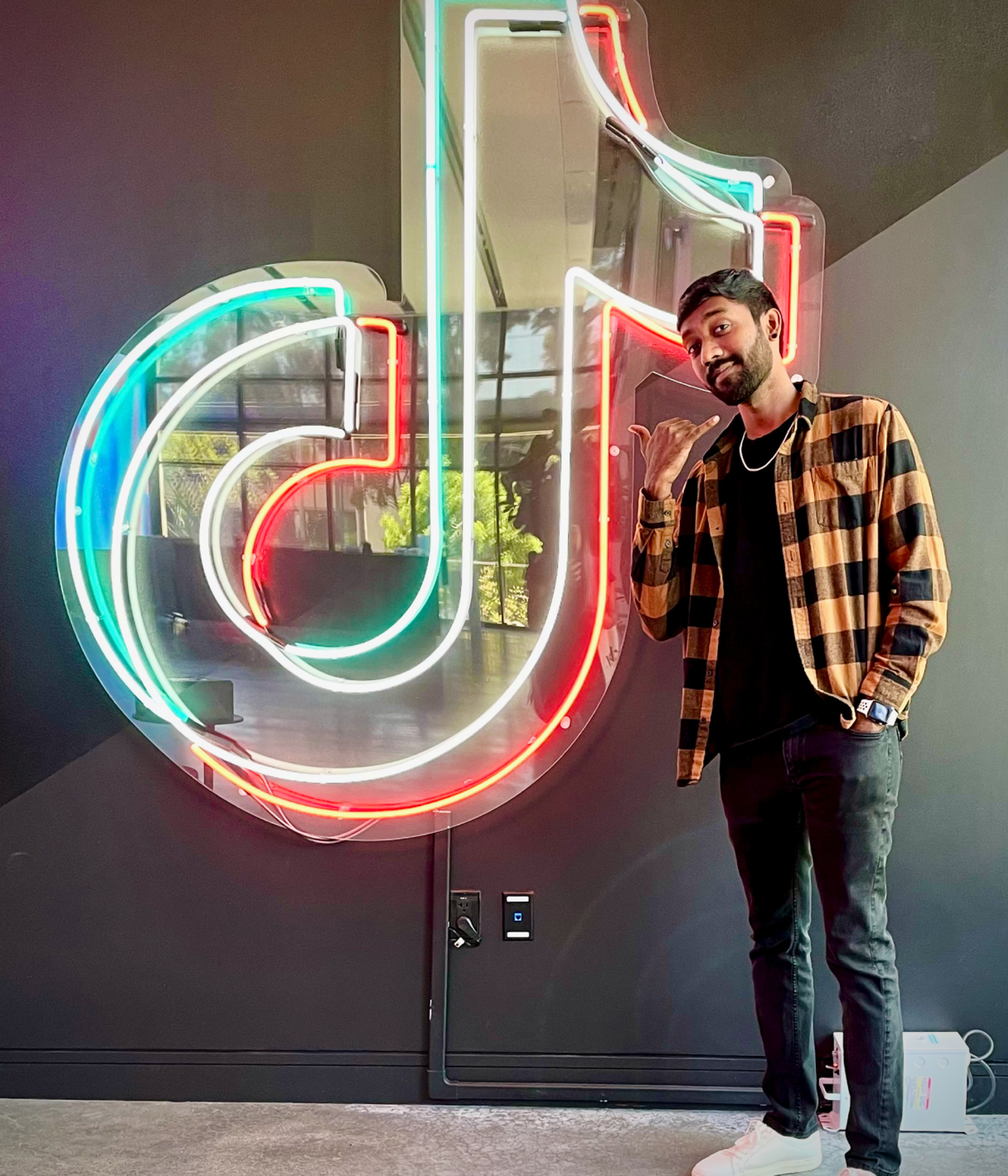 A young male TikTok employee with a beard and a plaid shirt poses in front of a neon sign of the TikTok music note.