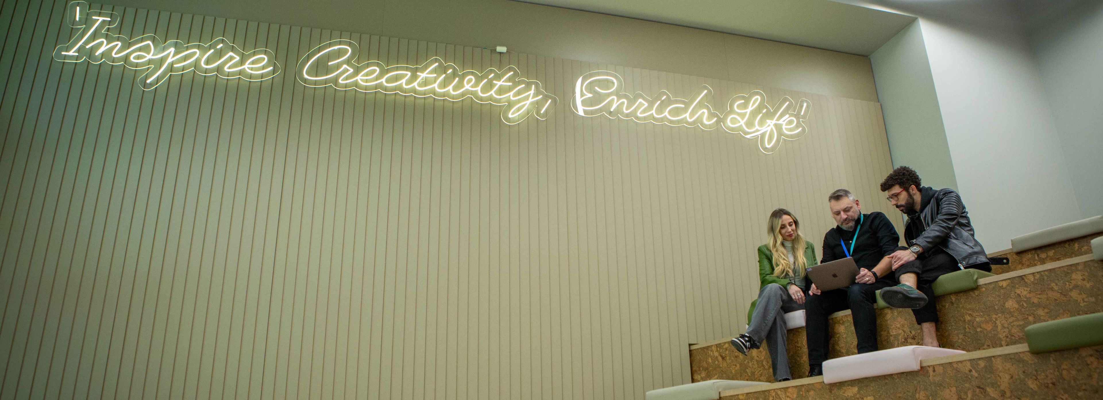 Three ByteDance employees sit on a set of stairs looking at a laptop and Inspire creativity, enrich life signage on the wall