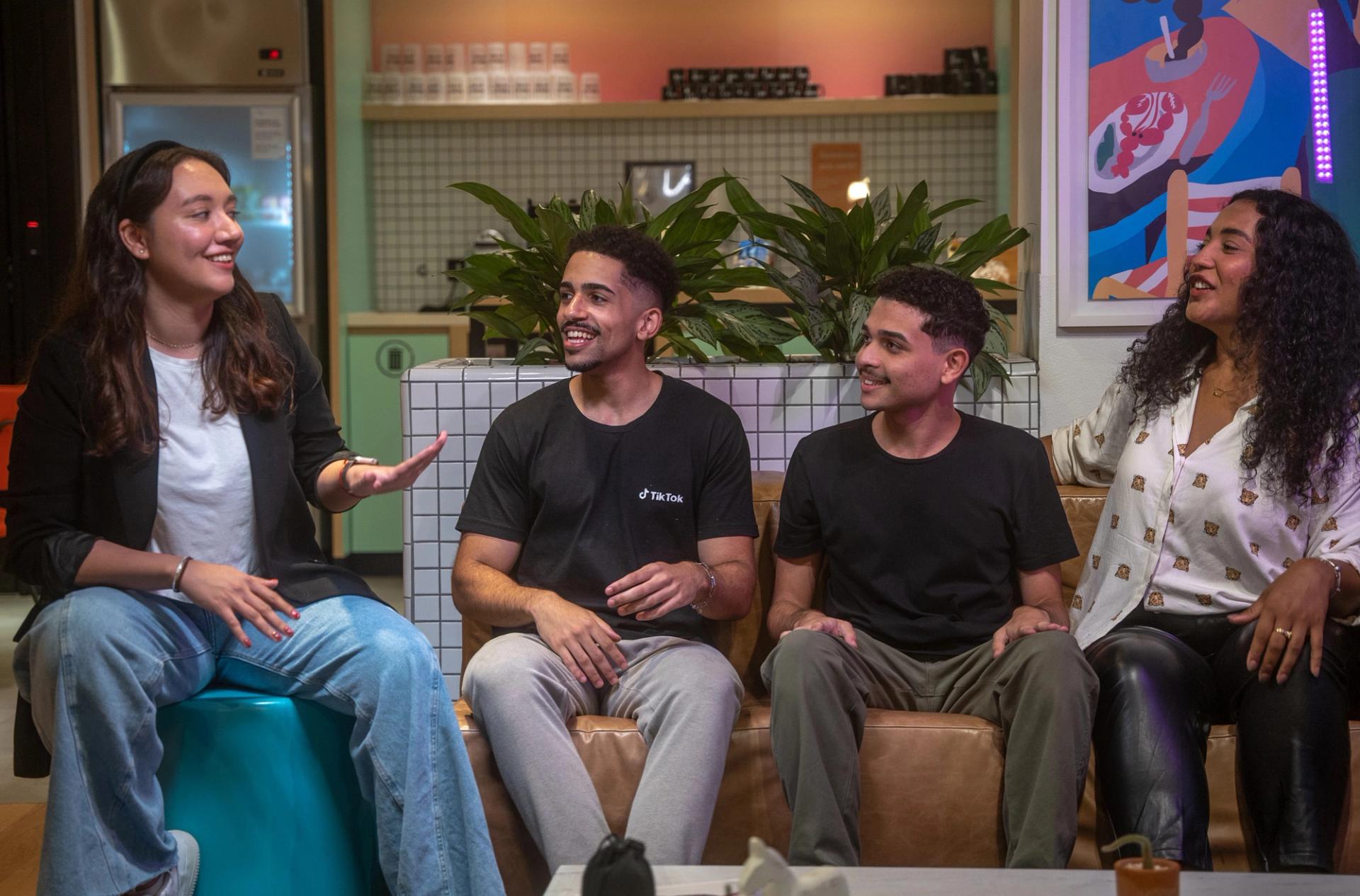 A diverse group of four young TikTok employees talk and smile as they sit beside each other on a couch.