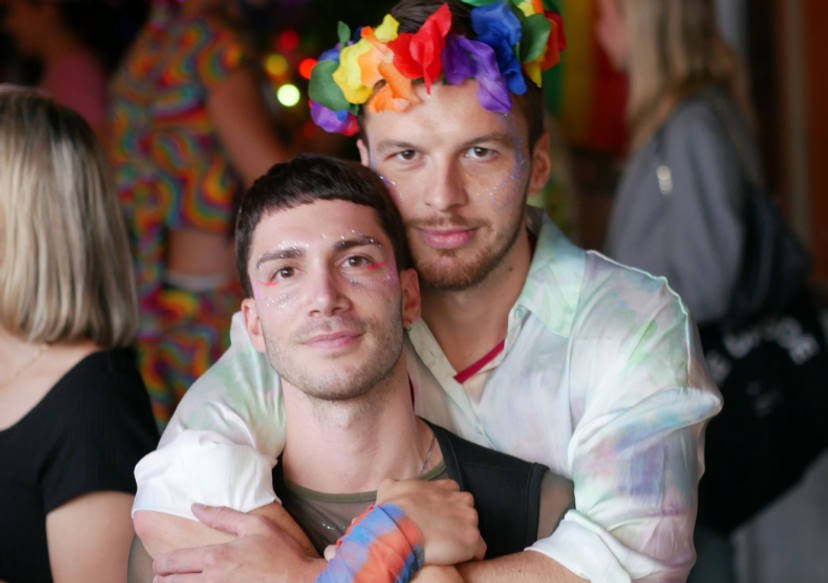 Two male TikTok employees wearing rainbow colored headdresses and shirts hug each other while looking at the camera.