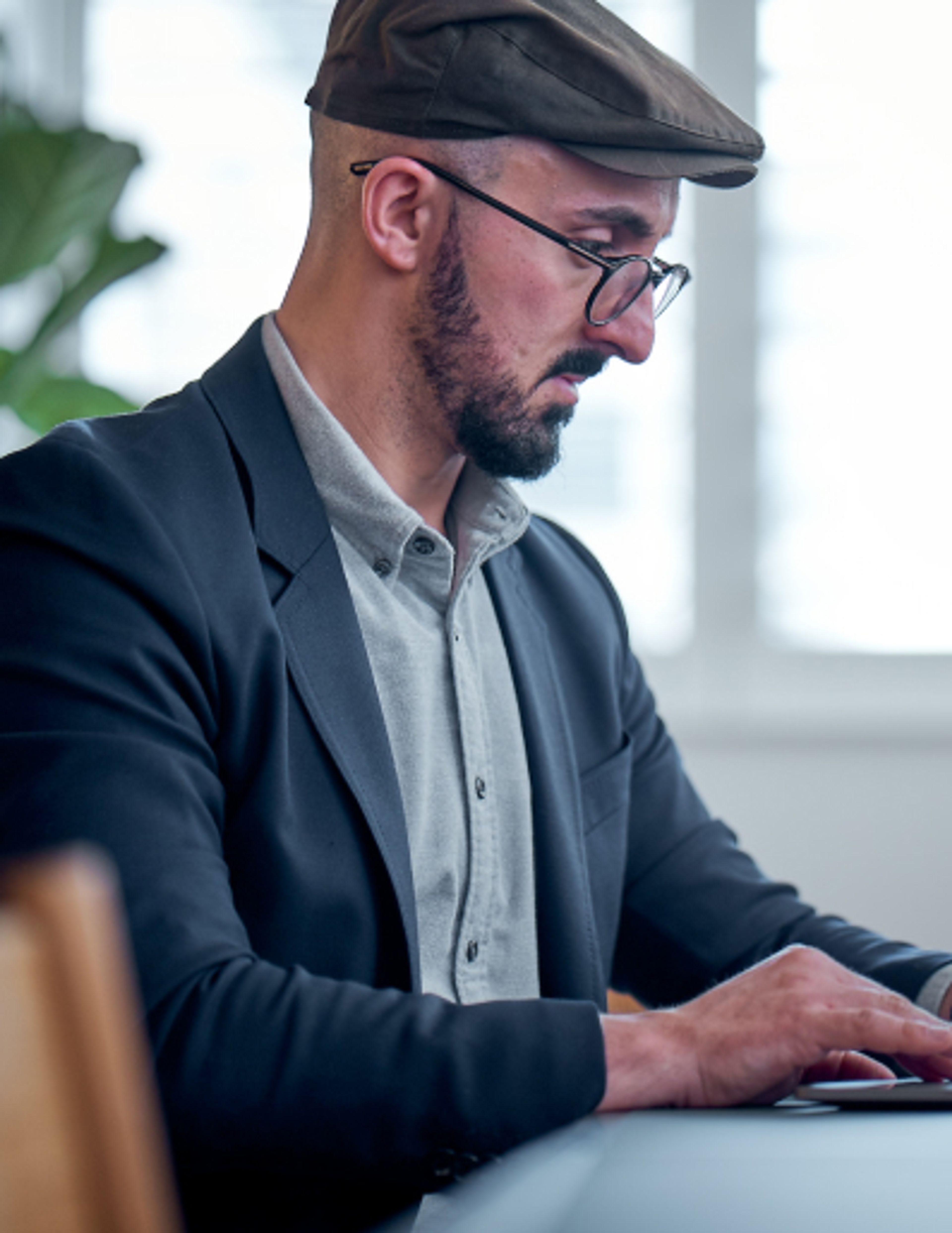 A bearded man with a cap and glasses works at his computer.