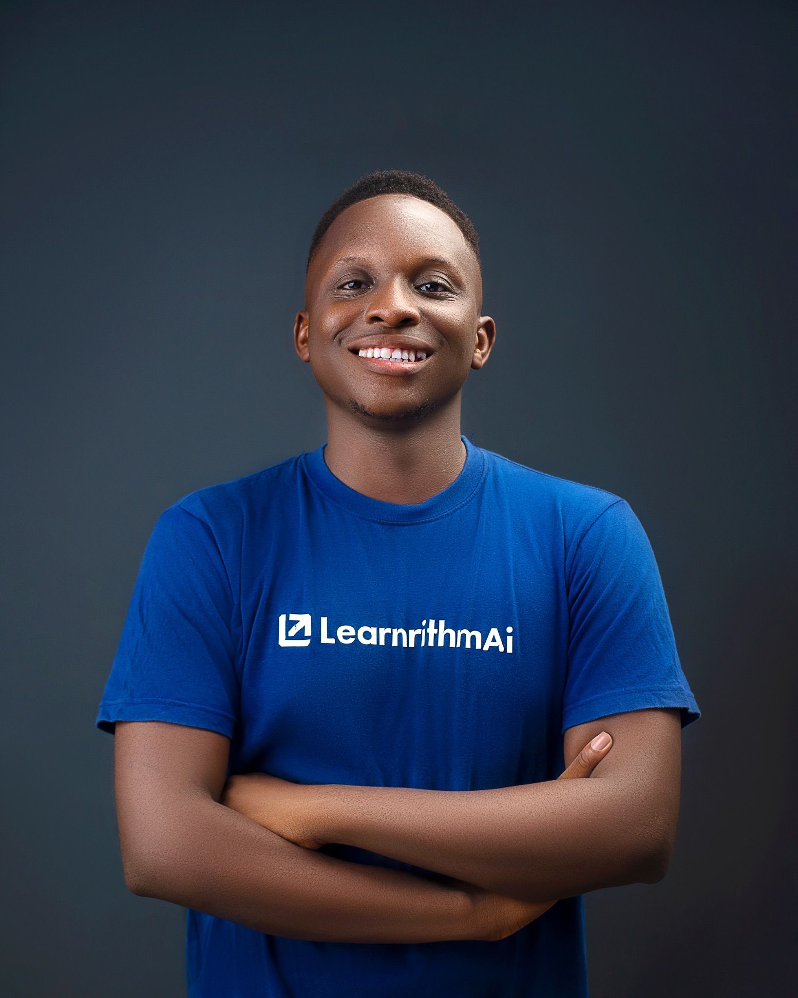 a man wearing a blue learnrithmai t-shirt is smiling with his arms crossed .pete