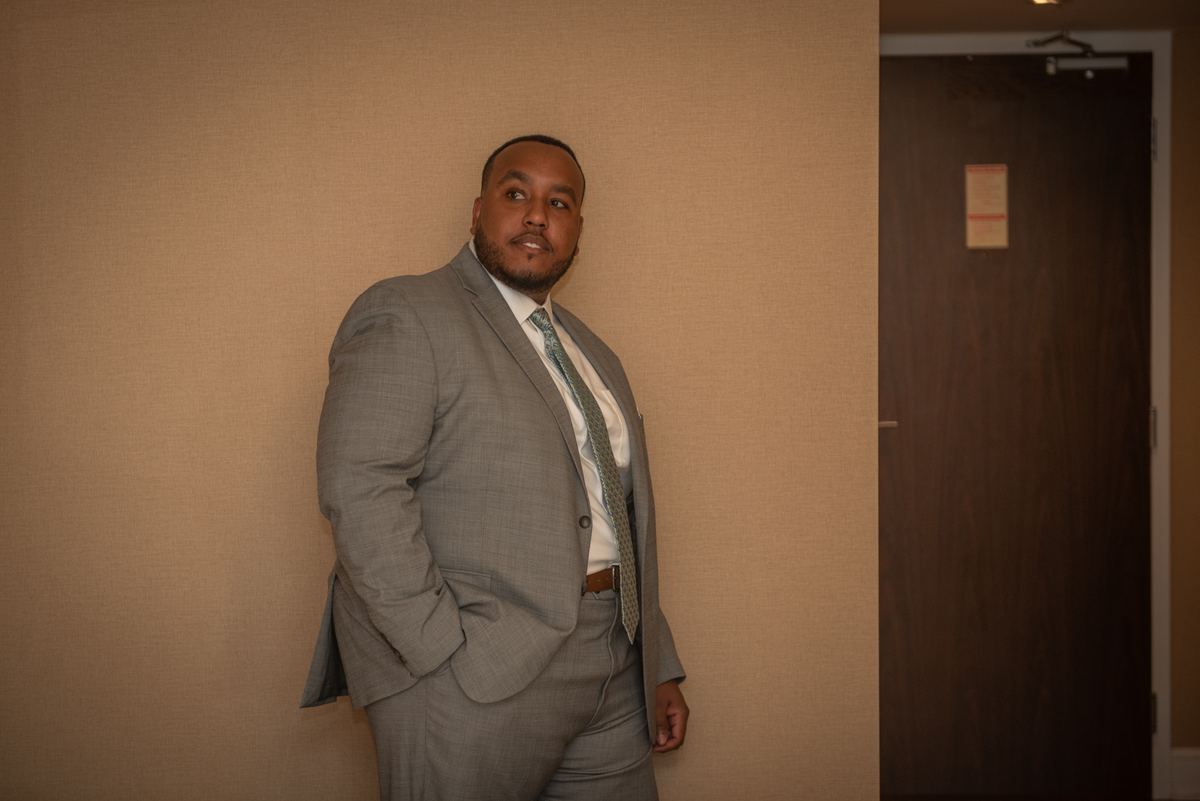 A man in a gray suit leans against a wall, looking thoughtfully upward in a warmly lit room with a dark wooden door nearby.