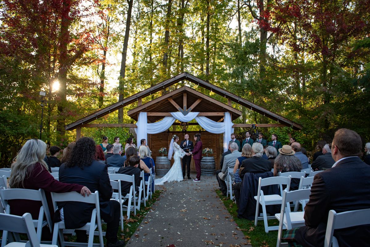 A bride and groom exchange vows under a wooden arbor surrounded by guests in white chairs, with autumn leaves on the ground.
