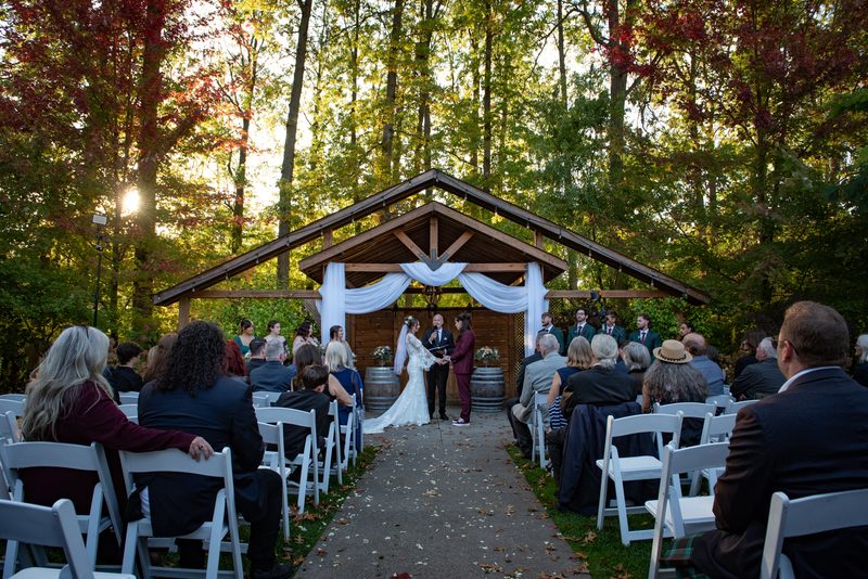 A bride and groom exchange vows under a wooden arbor surrounded by guests in white chairs, with autumn leaves on the ground.