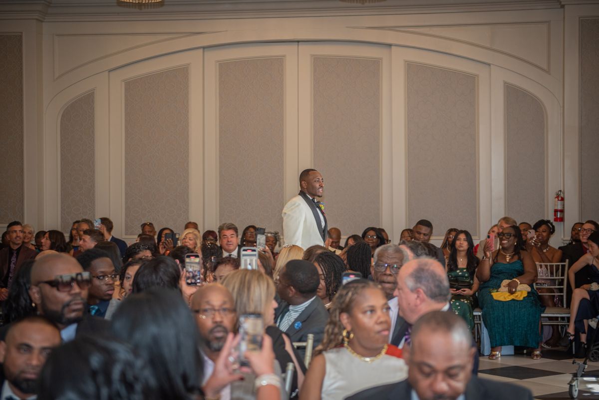 A man in a white tuxedo addresses an audience holding phones in a warmly lit banquet hall with ornate walls.