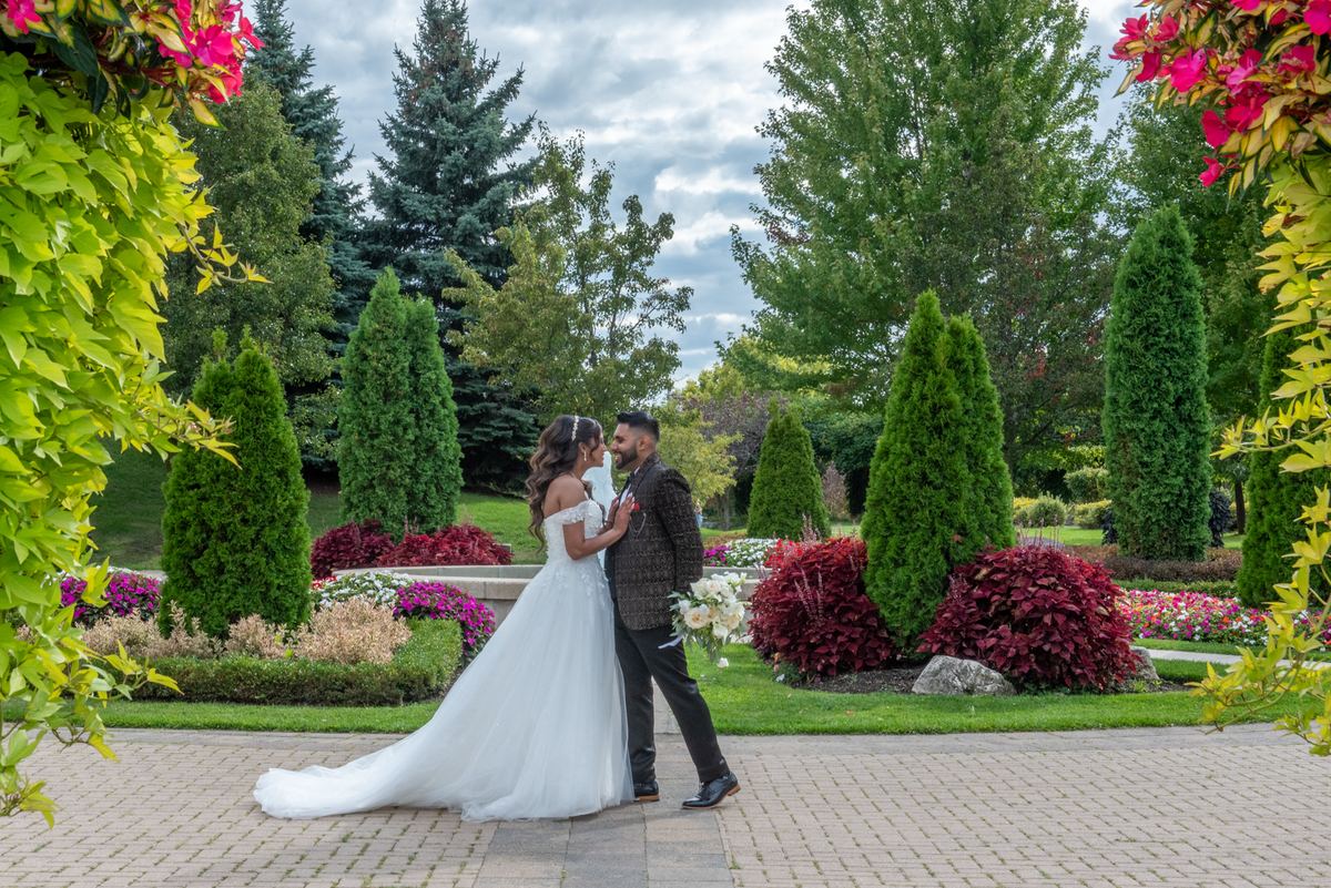 Bride and groom stand close on a garden path, gazing at each other with greenery and pink blossoms around them.
