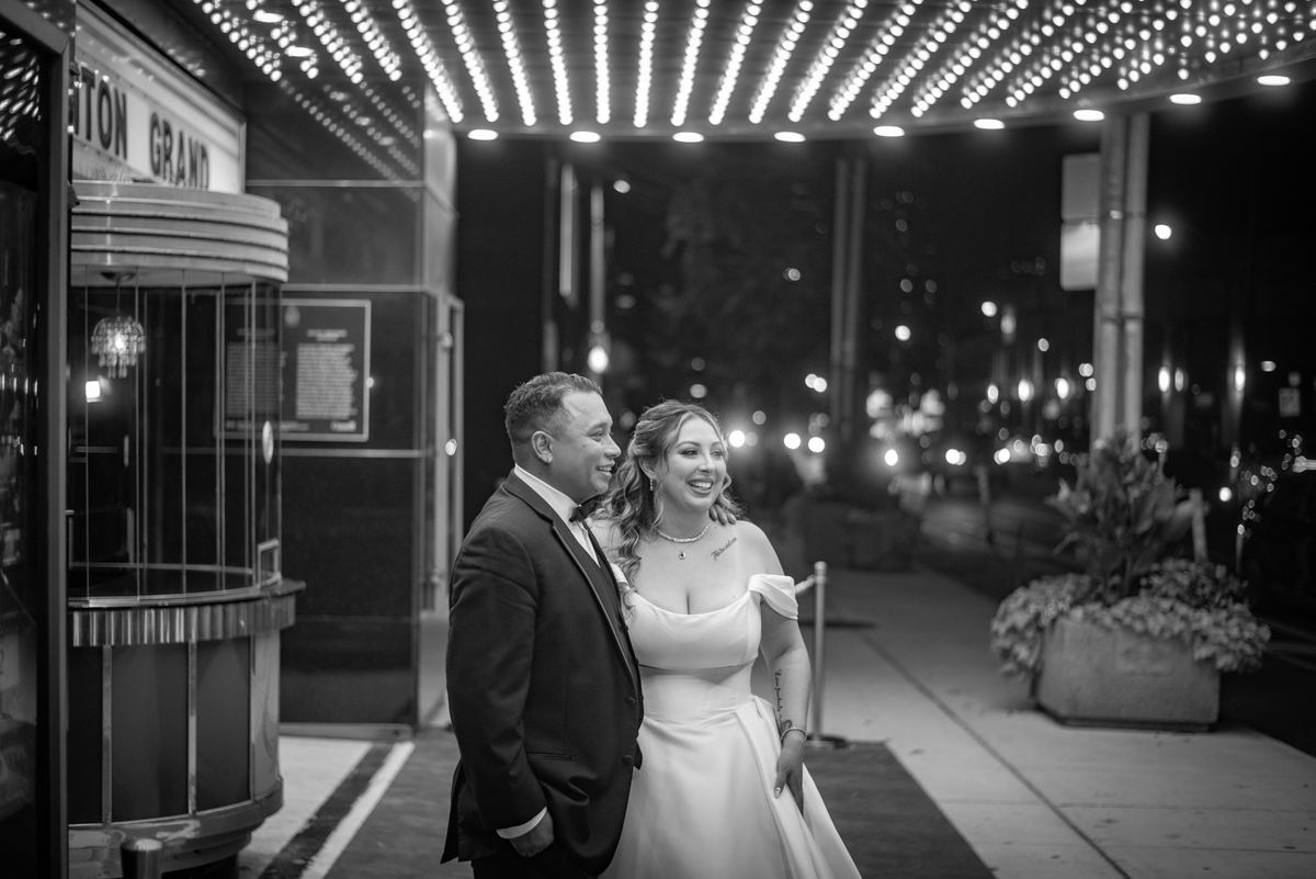 A smiling newlywed couple stands under the starburst lights of a theater entrance at night, illuminated by city glow.