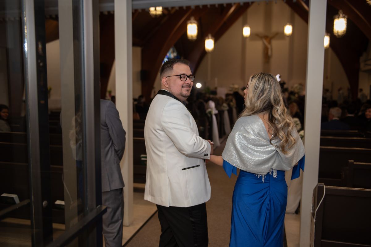 A man in a white tuxedo and a woman in a blue gown stand near church pews, illuminated by hanging fixtures.