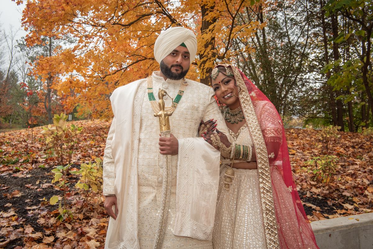A Sikh groom in white sherwani and turban with kirpan stands beside his bride in red and gold lehenga.