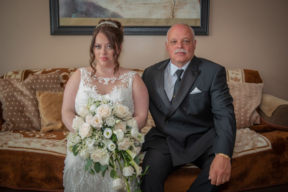 A bride in a white lace gown sits with her father on a sofa during a posed wedding portrait session.
