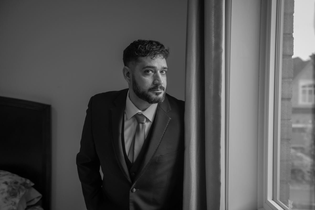 A man in a suit stands by a window, gazing thoughtfully into the distance, illuminated by soft natural light.