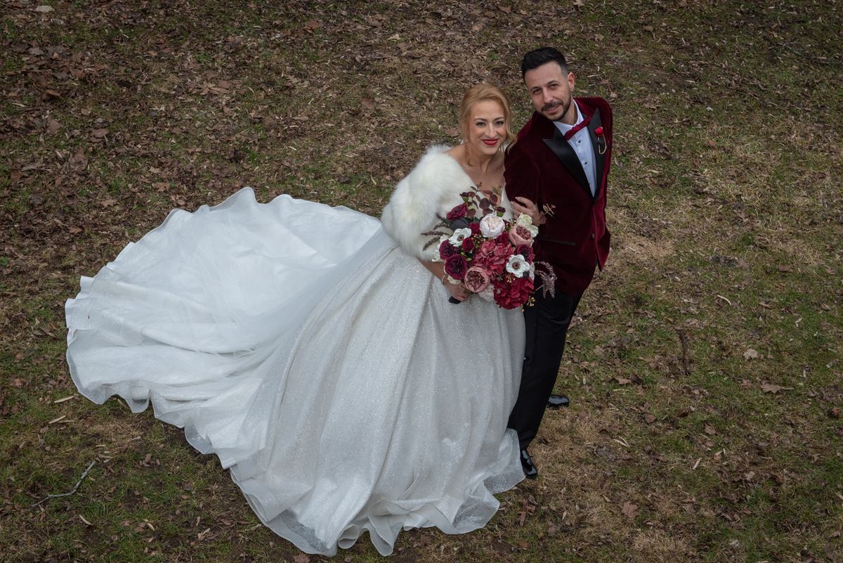 Bride in white gown and fur stole with groom in velvet tuxedo stand smiling on grassy field with fallen leaves.