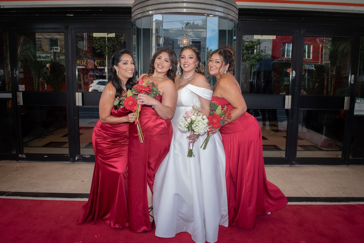 A bride and three bridesmaids pose outside a building's glass doors with bouquets of red gerbera daisies and white blooms.