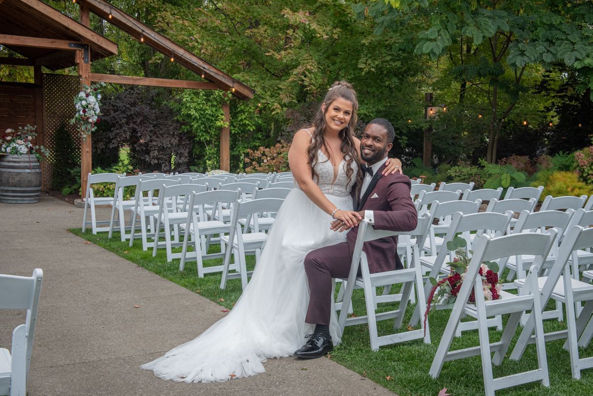A smiling bride and groom pose affectionately in an outdoor wedding aisle surrounded by greenery and chairs.