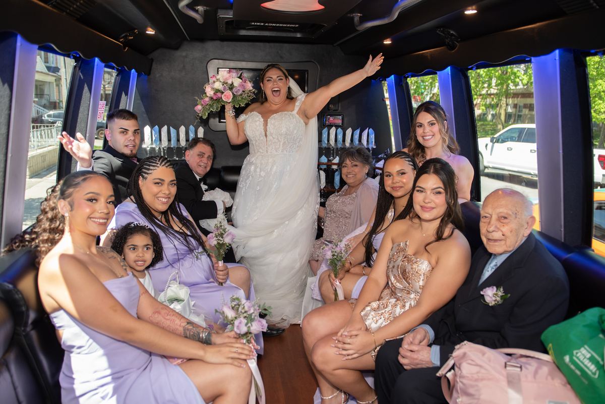 A smiling bride in a wedding dress waves with raised arms inside a festive limousine filled with family and bridesmaids.