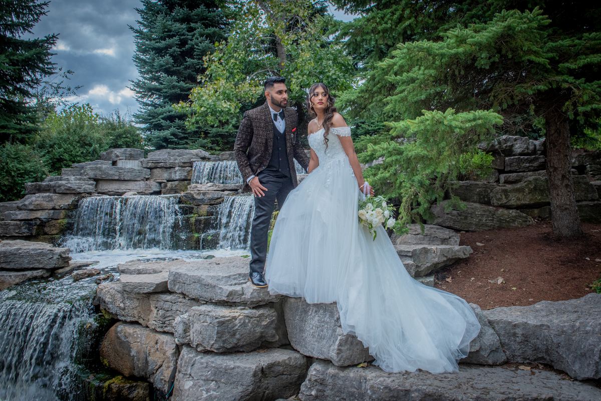 Bride in white gown and groom in suit stand by waterfall with evergreens behind them, gazing at camera.