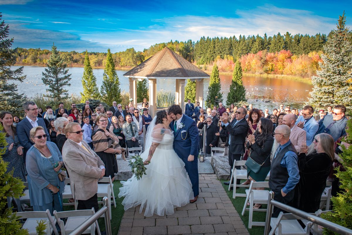 A newlywed couple kisses passionately as guests applaud beside a tranquil lake with autumn foliage and a wooden gazebo.