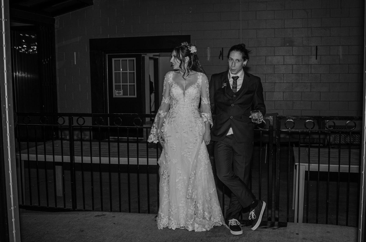 A newlywed couple walks at night, the bride in a lace gown and groom in suit with sneakers, under ambient light.