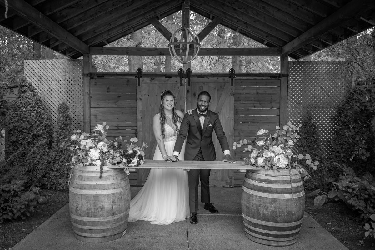 A newlywed couple smiles beneath a wooden pergola, holding a plank across wine barrels adorned with flowers.