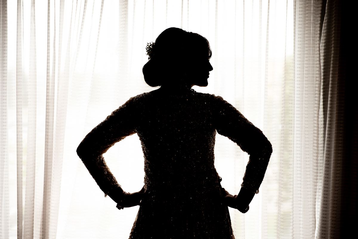 A woman in a sparkling garment stands confidently against a sheer curtain, hands on hips, in dramatic backlighting.