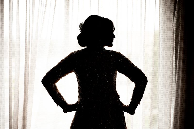 A woman in a sparkling garment stands confidently against a sheer curtain, hands on hips, in dramatic backlighting.