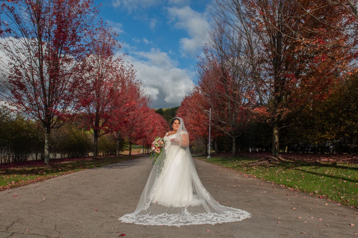 A bride in a white gown and long veil stands alone on a paved path framed by autumn trees, holding a bouquet.